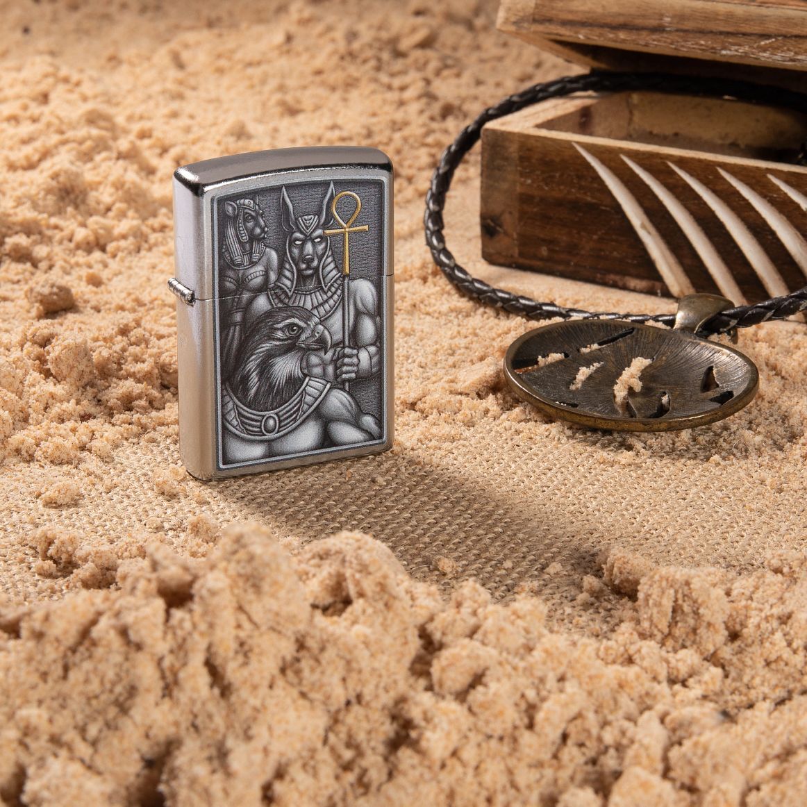 Lifestyle image of Egyptian Gods Design Street Chrome?äó Windproof Lighter standing in the sand with a box and necklace next to it
